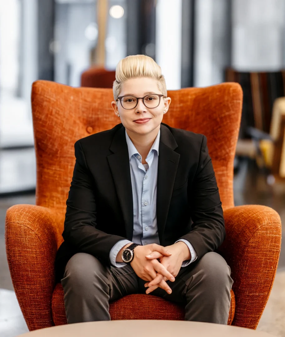 Professional portrait of a person wearing glasses and a blazer, seated on an orange armchair in a modern indoor setting
