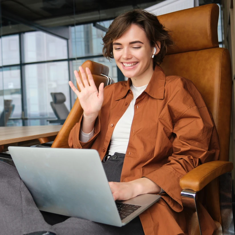 Woman sitting in an office chair, smiling and waving during a video call on her laptop